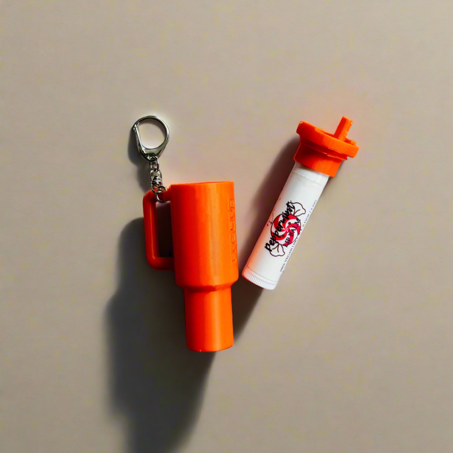 Orange silicone tumbler-style lip balm holder keychain with attached metal key ring displayed beside a matching orange lip balm tube on a neutral background.