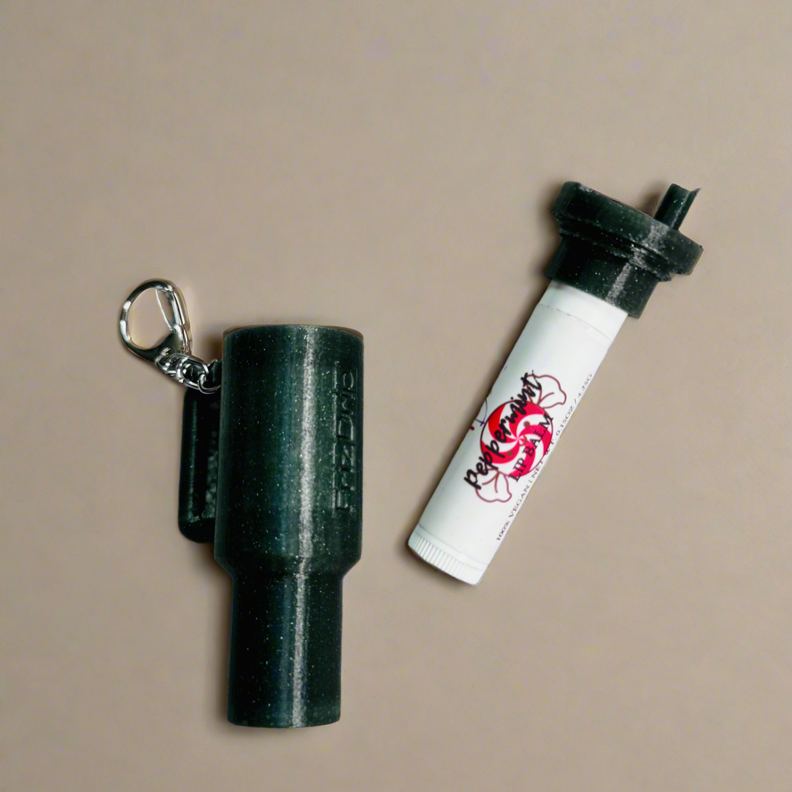 Black silicone tumbler-style lip balm holder keychain with detachable lid shown beside a standard lip balm tube on a beige background, designed to clip onto purses, backpacks, keys, and handbags.