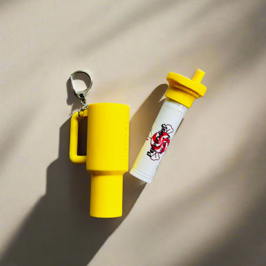 Yellow silicone tumbler-style lip balm holder keychain displayed beside a matching lip balm tube on a neutral background, featuring a durable bag charm with key ring for purses, backpacks, totes, and keys.