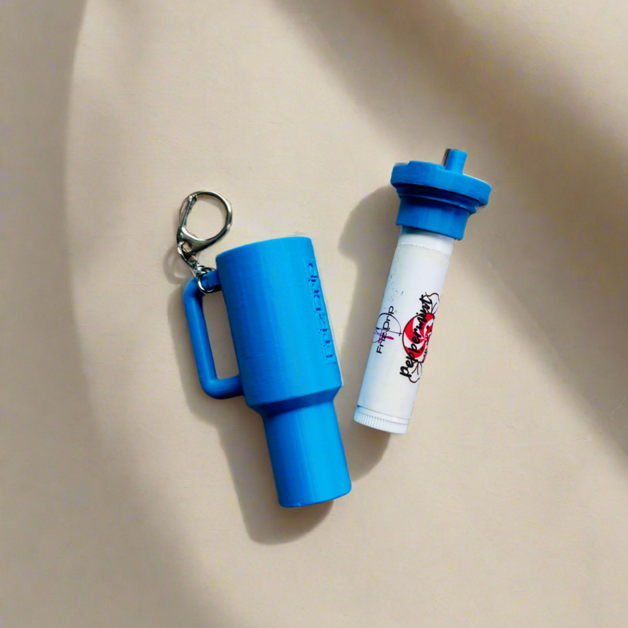 Blue silicone tumbler-style lip balm holder keychain displayed open beside a standard lip balm tube on a neutral beige background, showing the removable holder, keychain clip, and compact travel-friendly design.