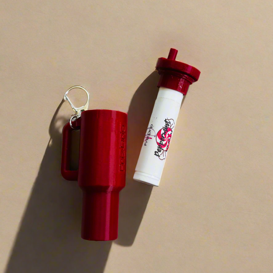 Red silicone tumbler-style lip balm holder keychain displayed beside a white lip balm tube with a matching red lid on a beige background, showing a portable bag charm for purses, backpacks, keys, and totes.