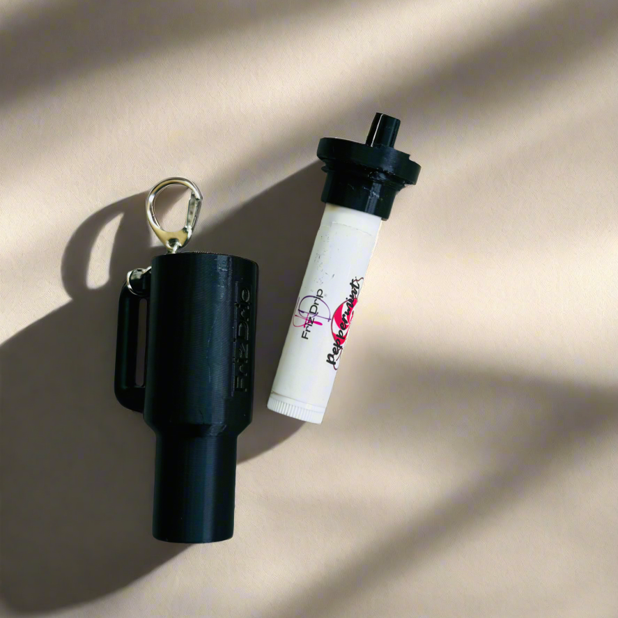 Black tumbler-style silicone lip balm holder keychain with a detachable key ring displayed beside a standard lip balm tube on a neutral background. Durable purse charm accessory designed to keep lip balm easily accessible on keys, handbags, backpacks, and totes.