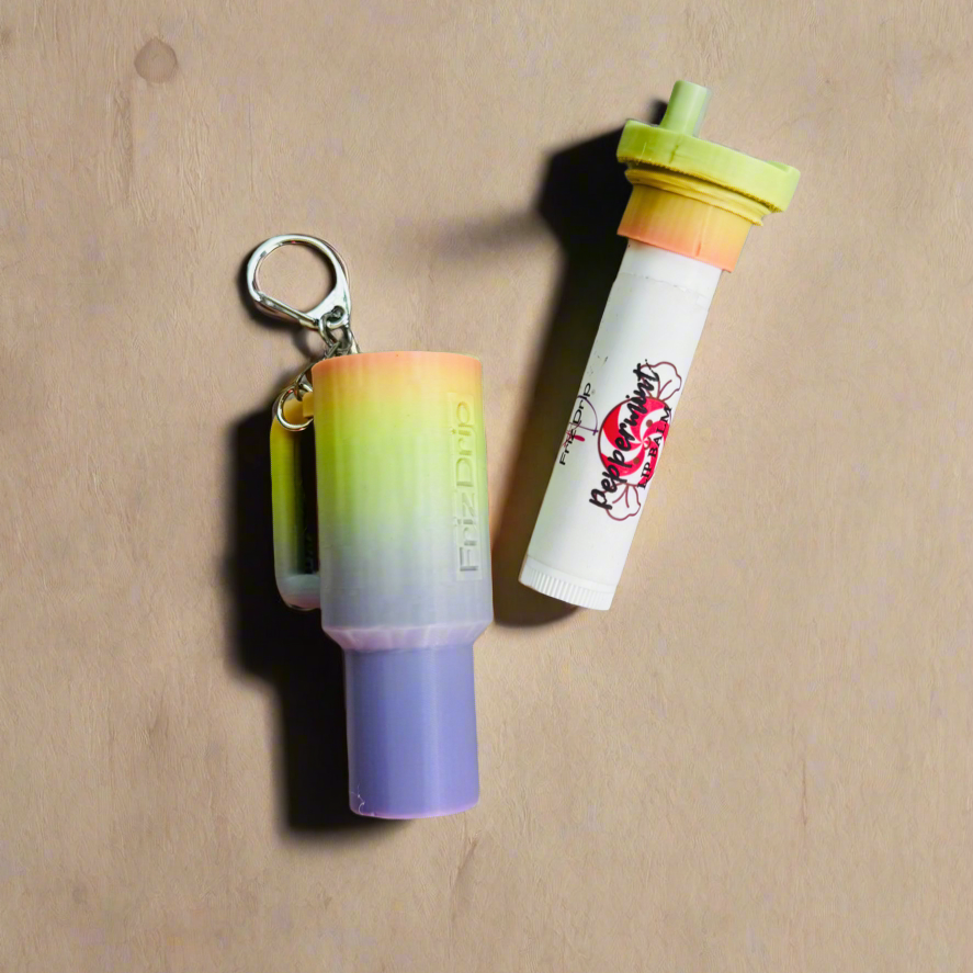 Rainbow gradient silicone tumbler-style lip balm holder keychain with silver clasp, displayed beside a standard lip balm tube on a neutral wood background. Reusable purse charm fits most standard lip balm tubes for backpacks, handbags, totes, and keys.