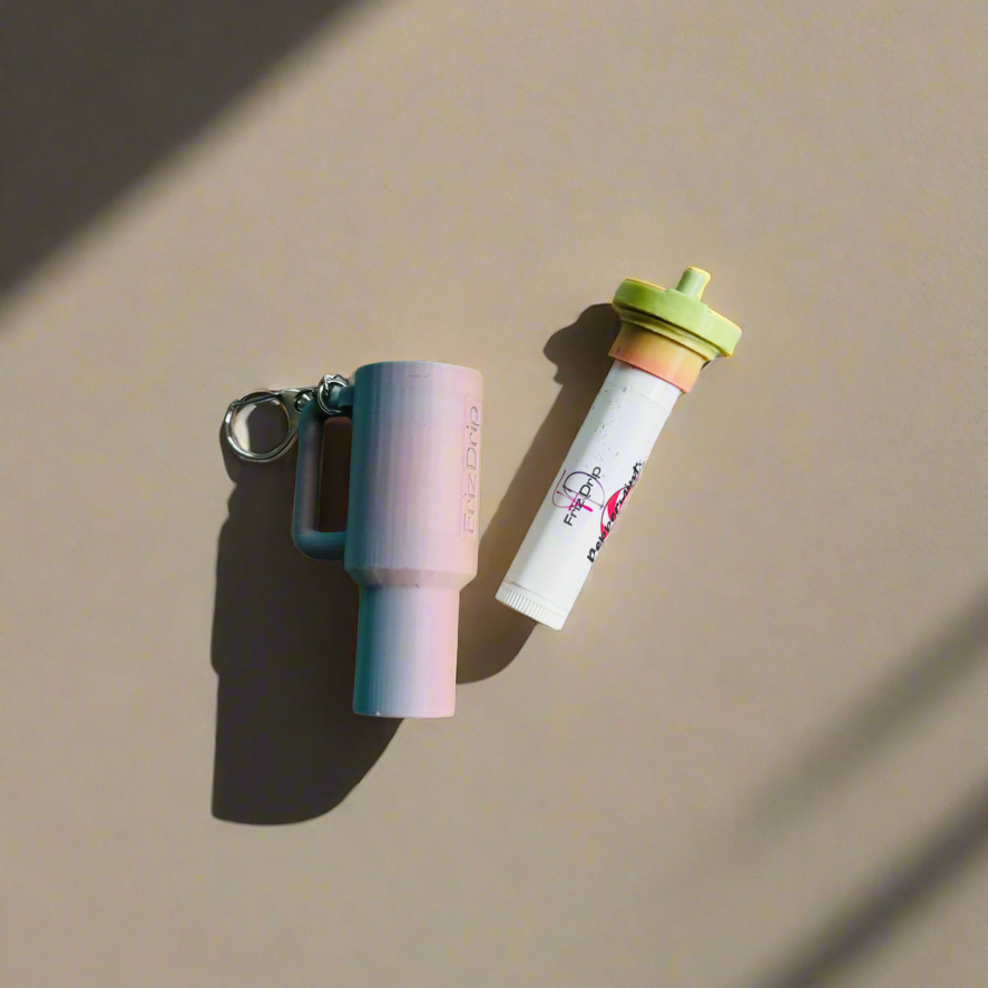 Pink tumbler-style silicone lip balm holder keychain displayed beside a standard lip balm tube on a neutral background, showing a cute bag charm accessory for purses, backpacks, and keys.