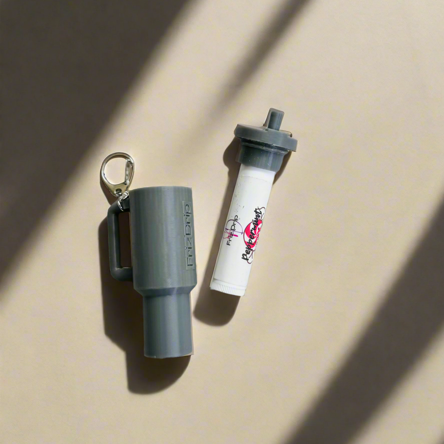 Gray silicone tumbler-style lip balm holder keychain displayed beside a standard lip balm tube on a neutral background, showcasing a reusable key ring clip for attaching to purses, backpacks, totes, or keys.