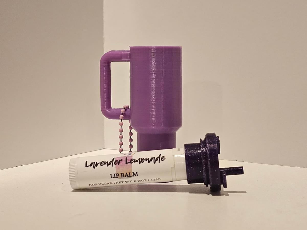 Purple tumbler-style silicone lip balm holder keychain displayed with a lavender lemonade lip balm tube and detachable key ring, designed to clip onto purses, backpacks, keys, and handbags for convenient everyday carry.