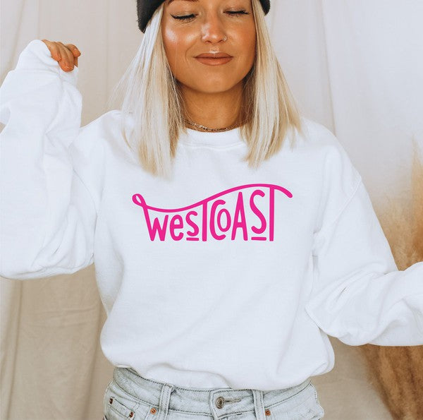 Woman wearing a white West Coast graphic crewneck sweatshirt with bright pink lettering, styled with light wash denim jeans and a black beanie in a cozy indoor lifestyle setting.