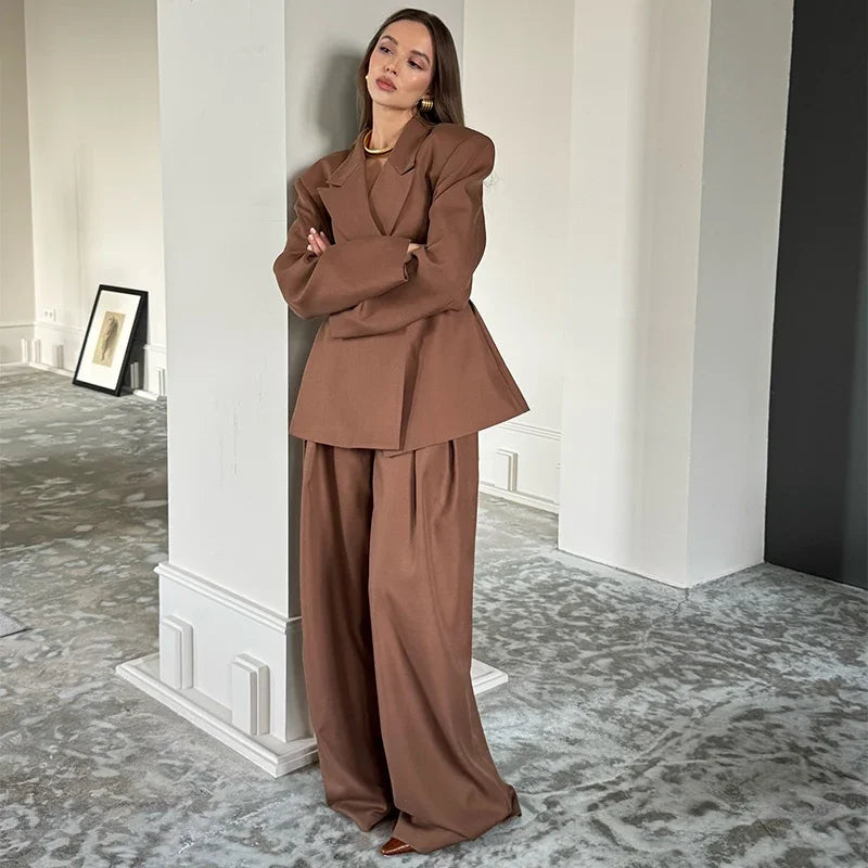 Woman in a brown suit standing in an elegant room with white walls and a patterned rug.