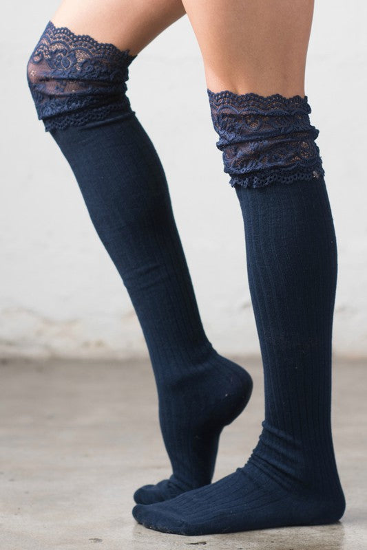 Woman wearing dark blue ribbed over-the-knee socks featuring elegant floral lace top detailing. These chic thigh-high socks offer a soft stretch fit and versatile styling for layering with dresses, skirts, boots, oversized sweaters, and cozy seasonal outfits. Designed for comfort and fashion, the textured knit construction and feminine lace trim create a sophisticated look perfect for fall, winter, cottagecore fashion, casual wear, lounging, and everyday elegance. Women's over-the-knee socks with lace a