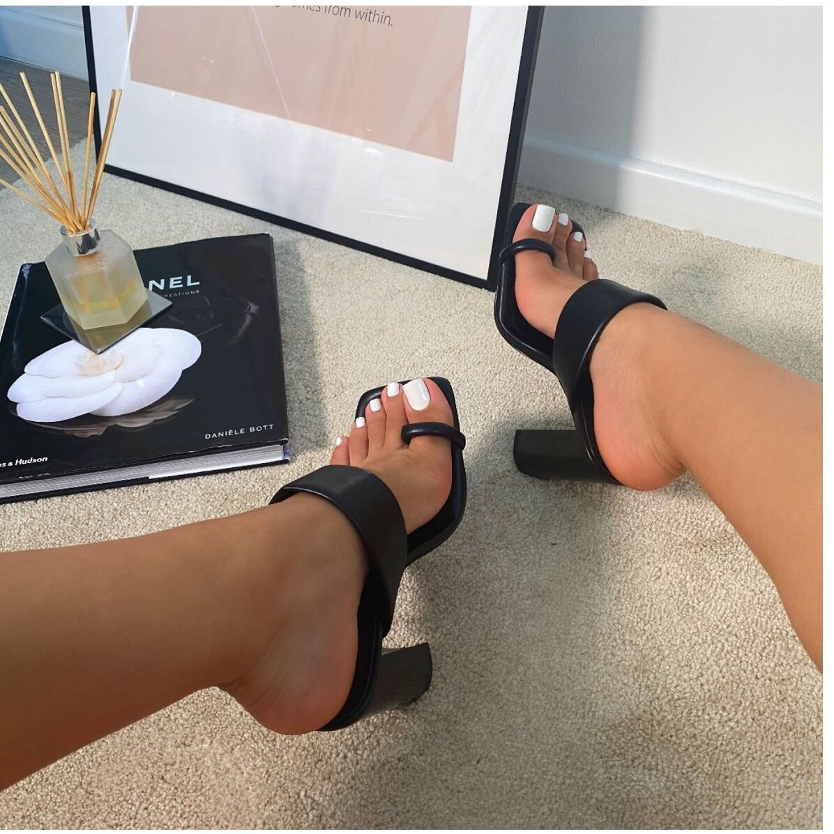 Black sandals worn by a person on a carpeted floor with a Chanel book and diffuser in the background.