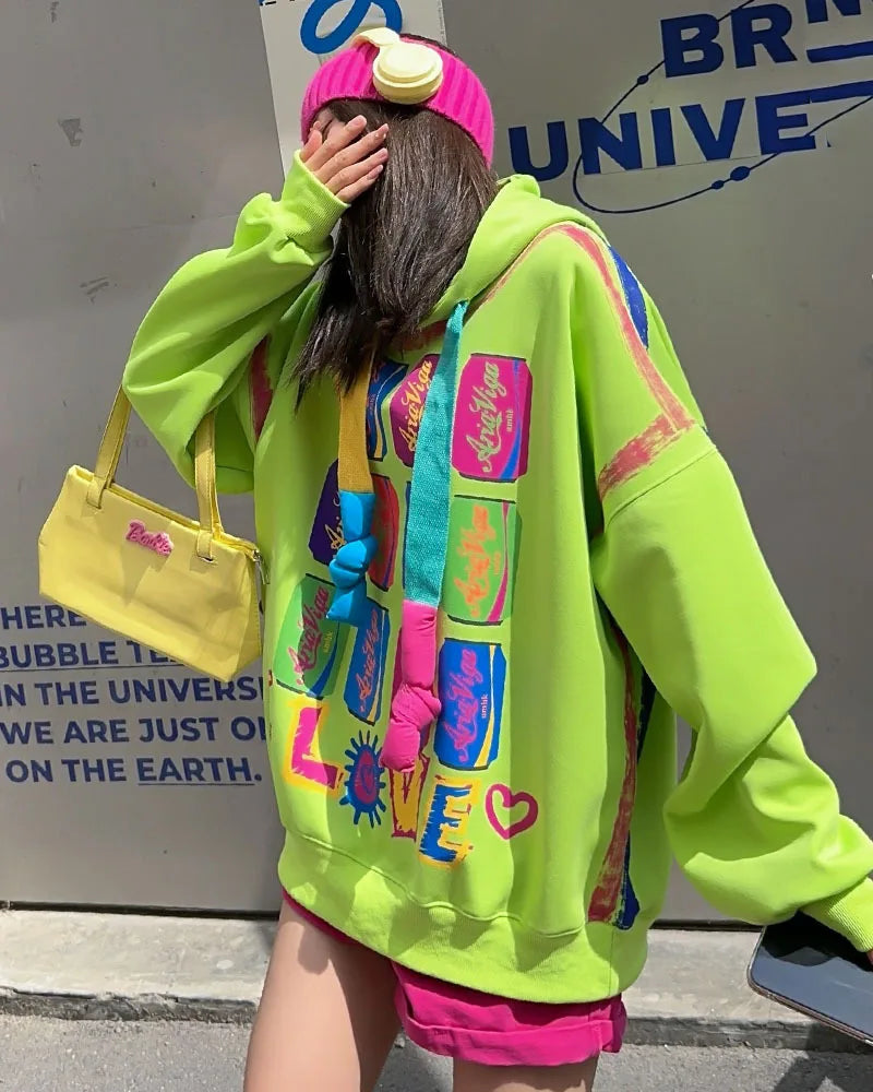 Person wearing a bright green hoodie with colorful designs, holding a yellow bag, in front of a branded wall.