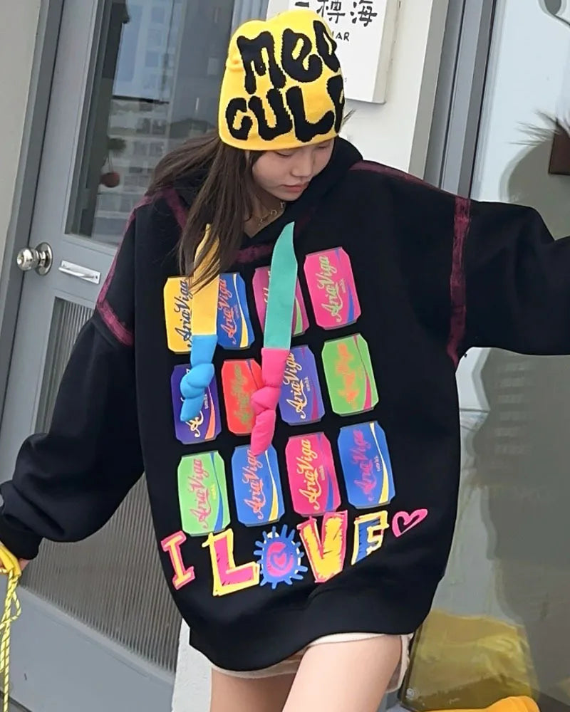 Person wearing a colorful hoodie with text and a yellow hat with text, standing indoors.