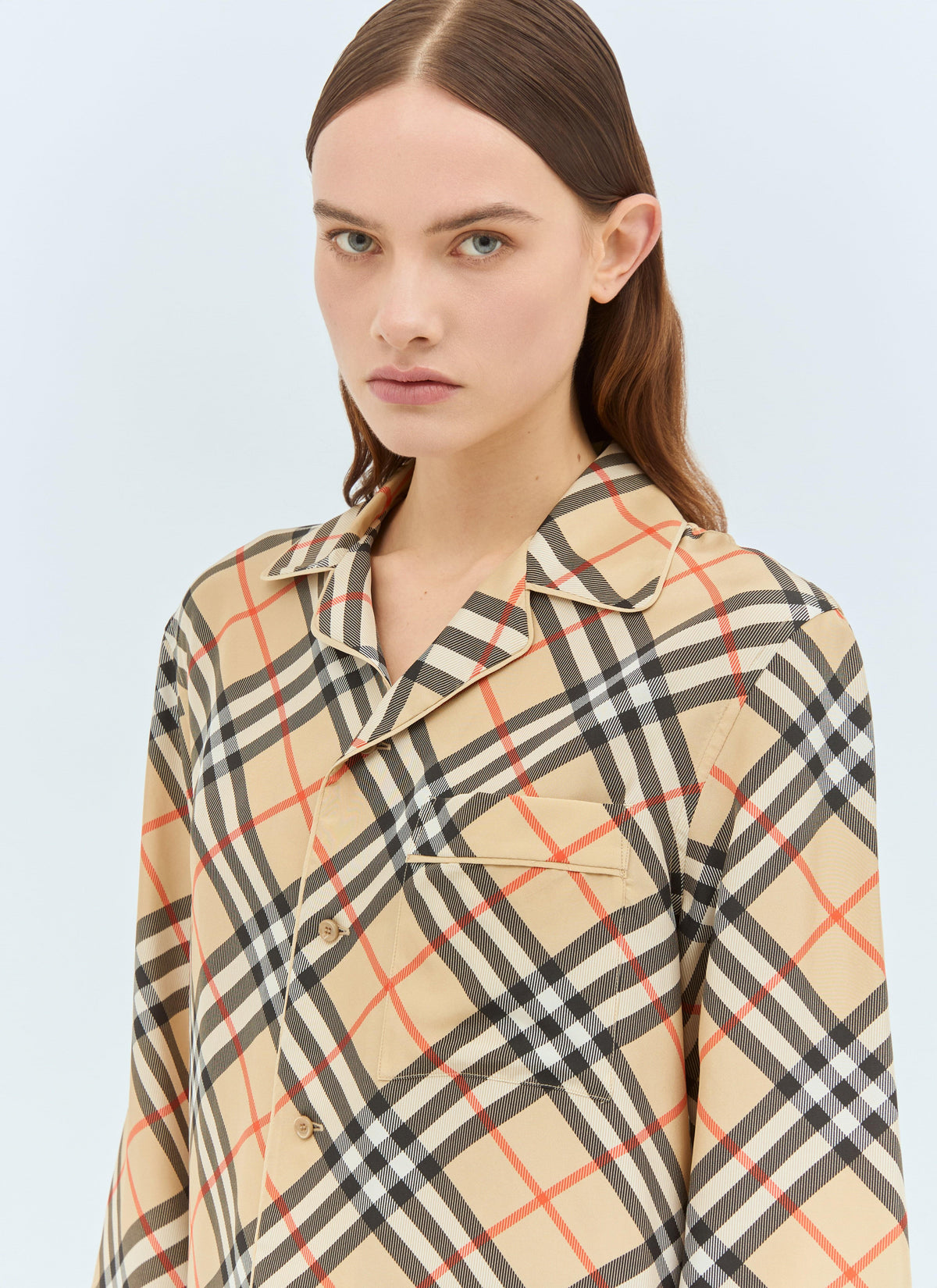 A woman wearing a Burberry check silk pyjama shirt with a revere collar and long sleeves, styled against a soft light blue background.
