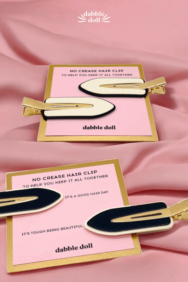 Two gold hair clips on pink cards with 'dabble doll' branding against a pink fabric background.