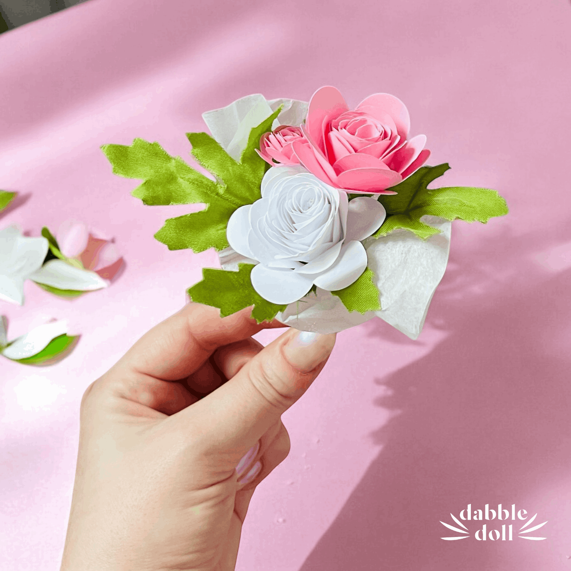 Hand holding a small bouquet of pink and white flowers with green leaves on a pink background, featuring the brand 'Dabble Doll'.