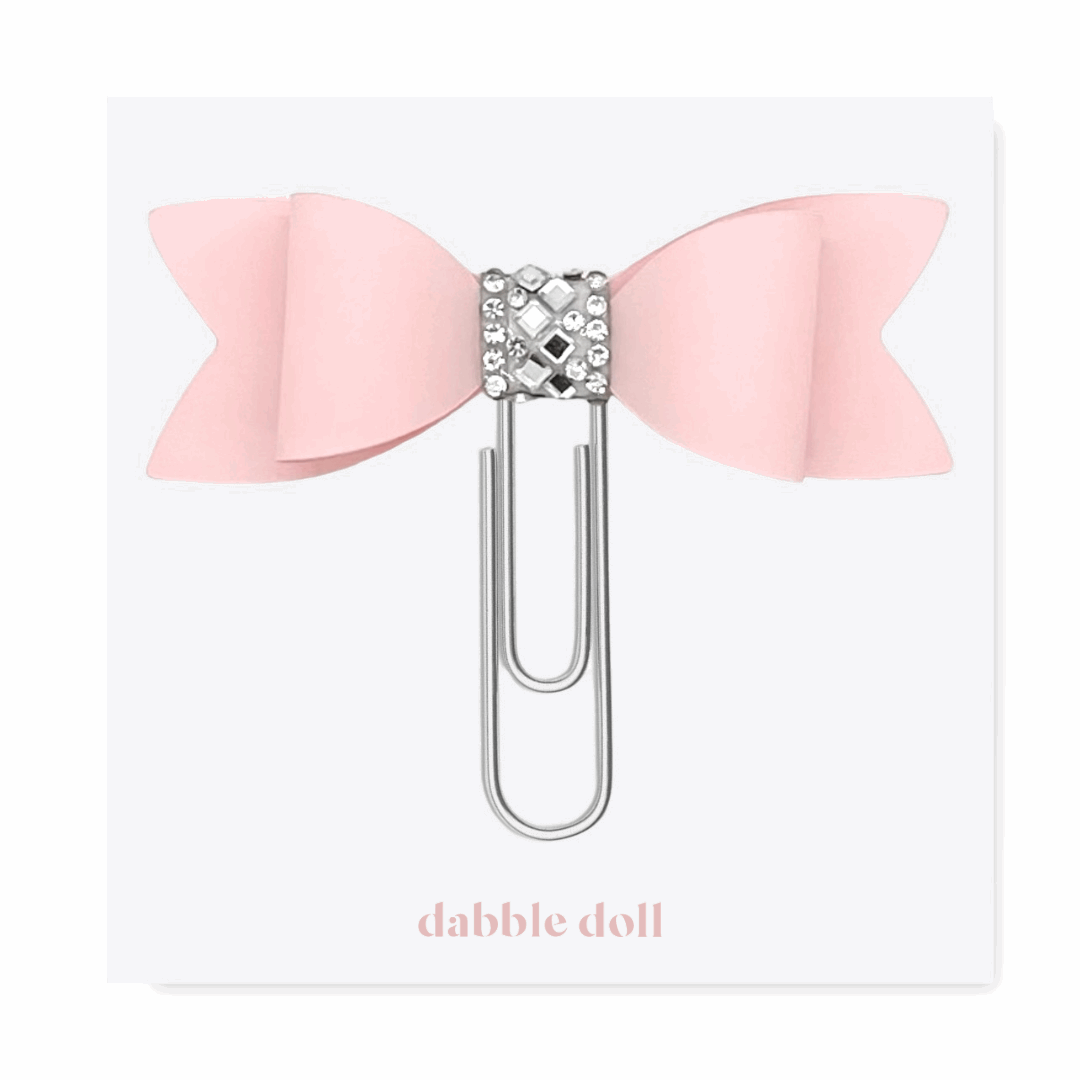 Pink bow-shaped paper clip with a decorative center on a white background, branded 'dabble doll'.