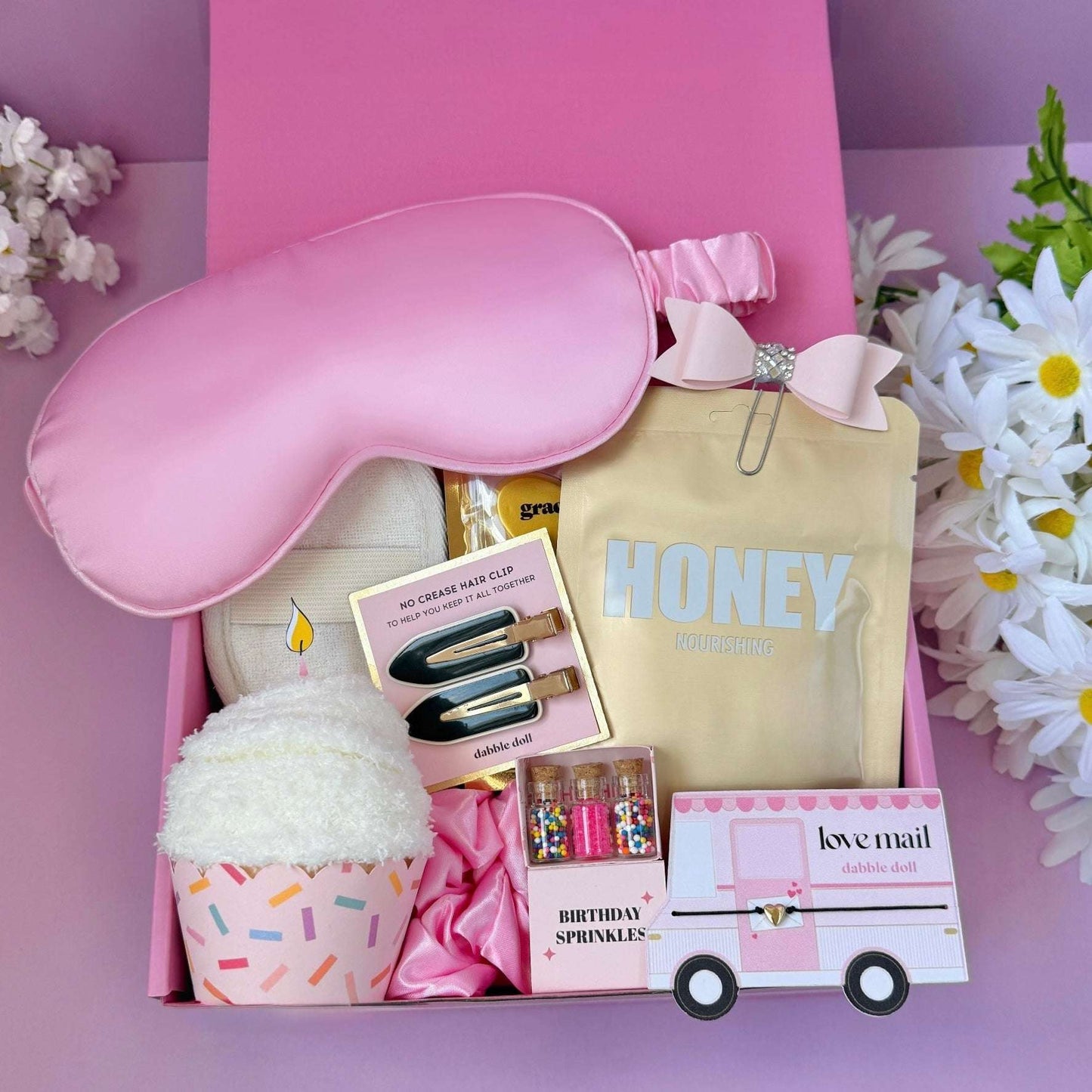 Pink gift box with sleep mask, hair clips, and other items on a pink background with flowers.