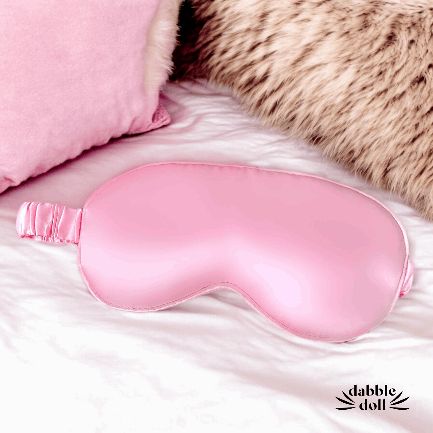 Pink satin eye mask on a soft surface with 'dabble doll' branding.