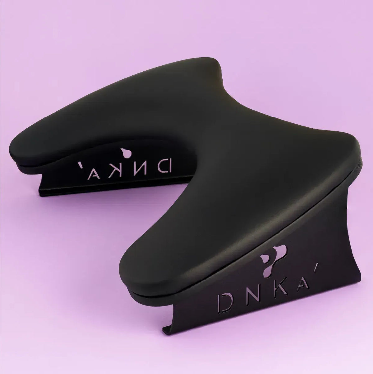 Anatomical black manicure armrest with the DNKa logo etched on the metal leg, displayed against a soft pink background.