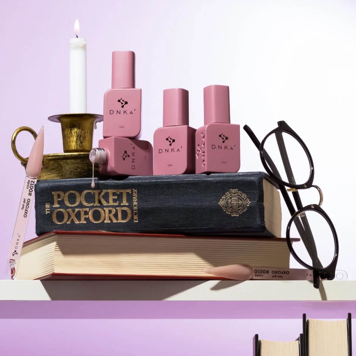 Pink DNK4 nail polish bottles arranged on top of stacked books, with a candle in a gold holder and black eyeglasses nearby, against a pale lavender background.
