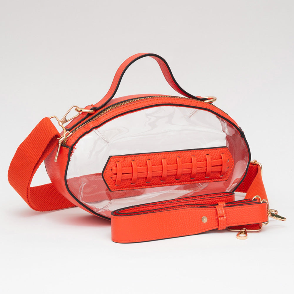 A clear oval handbag with bright orange trim, handle, zipper, and matching orange strap, featuring a faux leather football-style patch on the front and gold-tone hardware, photographed on a clean white background.