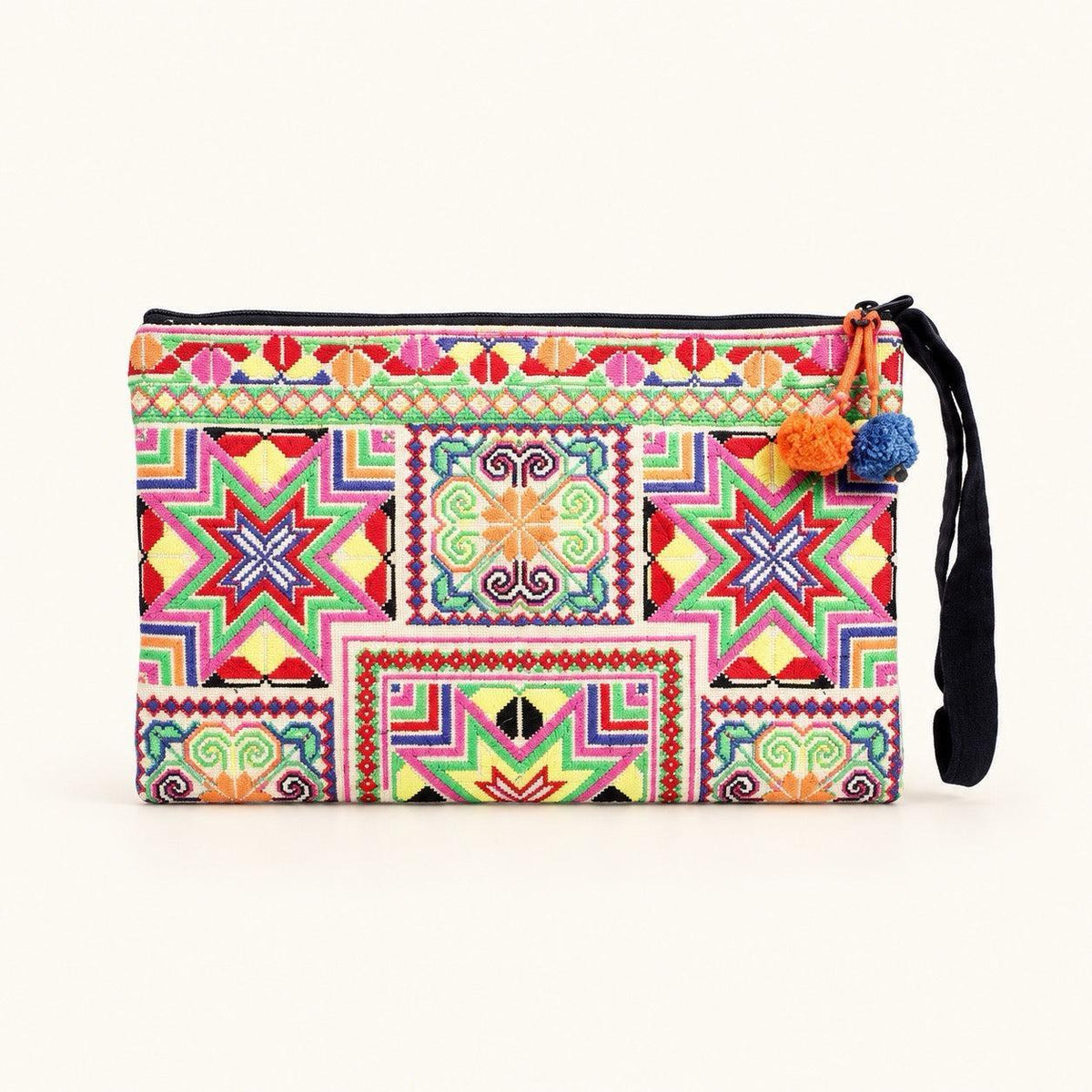 A rectangular wristlet clutch with a vibrant, multicolor geometric and tribal-inspired pattern, featuring a black zipper, a black wrist strap, and decorative pom-pom tassels on the side, set against a plain off-white background.