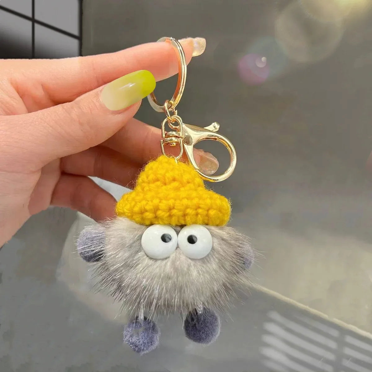 Keychain with a fluffy face and yellow hat held by a hand against a neutral background