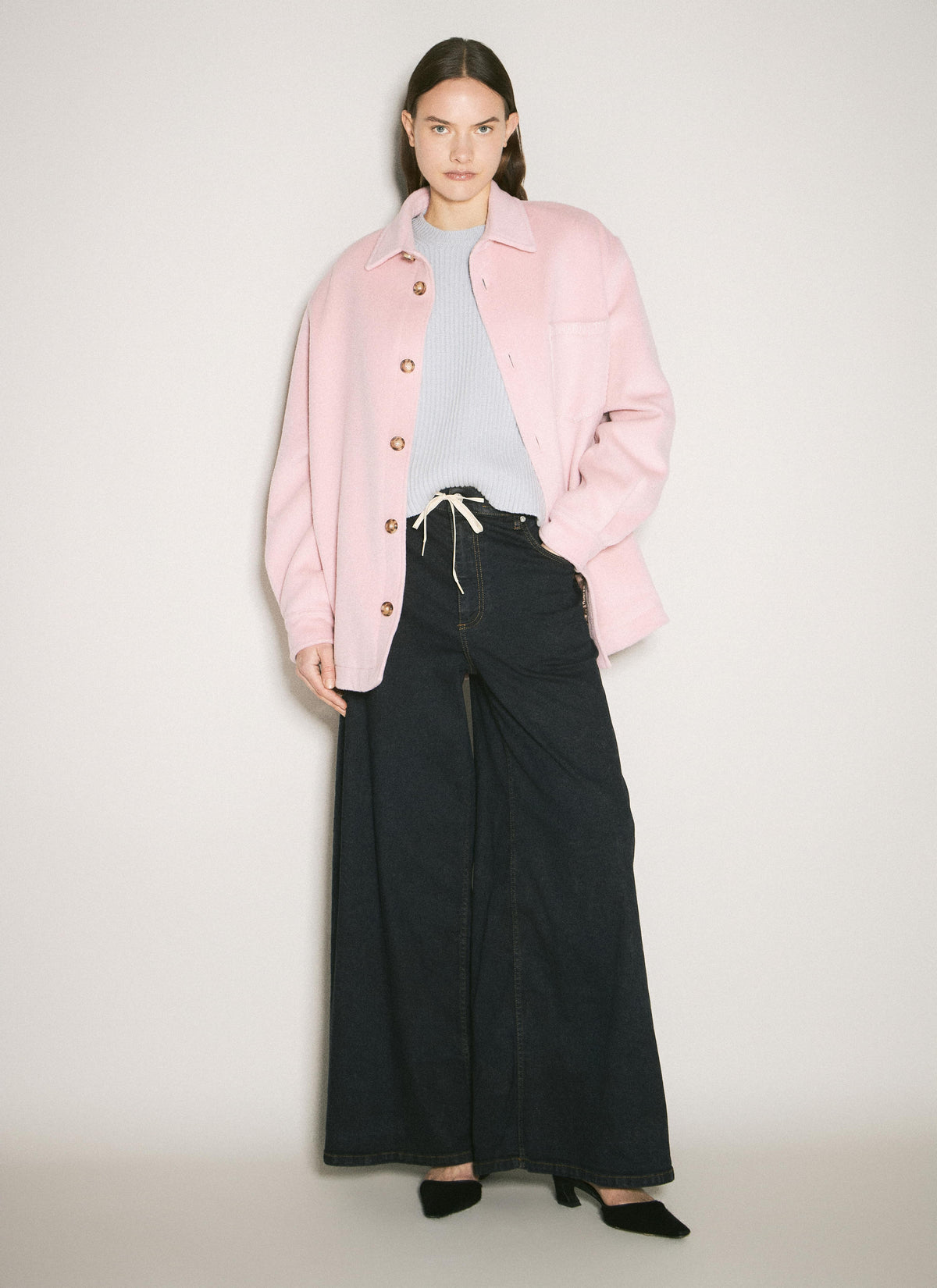 Elegant studio shot of a model wearing a pink jacket over a gray shirt and black trousers, set against a clean, minimalist background.