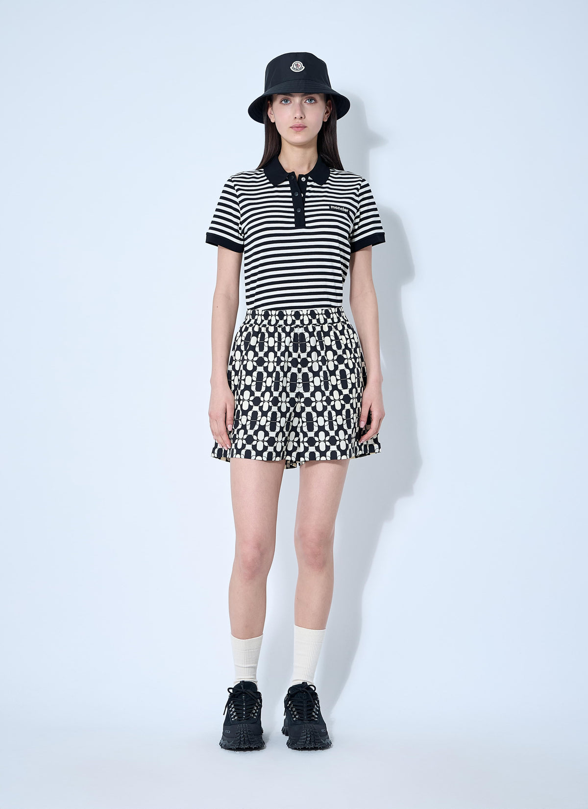 A model stands against a light blue studio background wearing a black and white horizontally striped short-sleeve polo shirt paired with a matching geometric patterned mini skirt, a black bucket hat, white socks, and black sneakers.