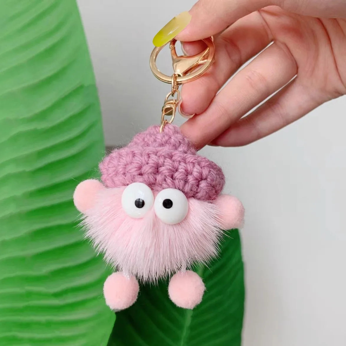 Hand holding a fluffy pink keychain with googly eyes against a green leaf background