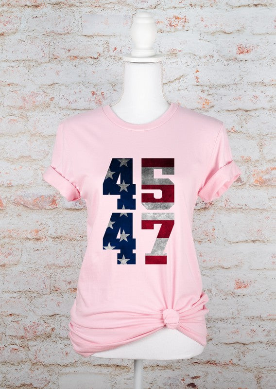 Pink unisex graphic T-shirt on a mannequin featuring a bold "45 47" patriotic American flag design, with rolled short sleeves and a knotted front hem, displayed against a white brick wall.