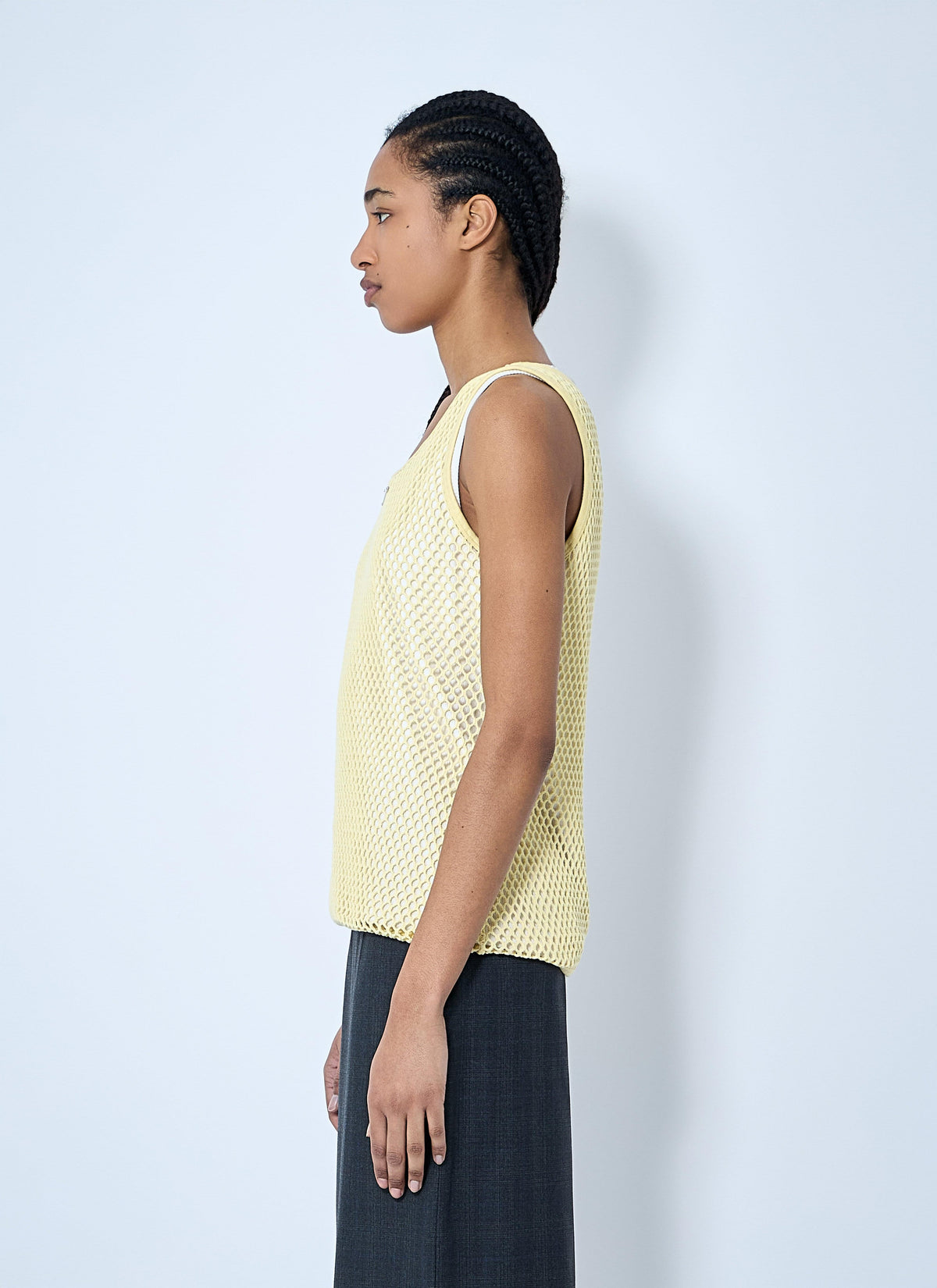 A model shown in profile wearing a pale yellow open-knit sleeveless top and dark charcoal gray wide-leg trousers, standing against a light gray studio background.