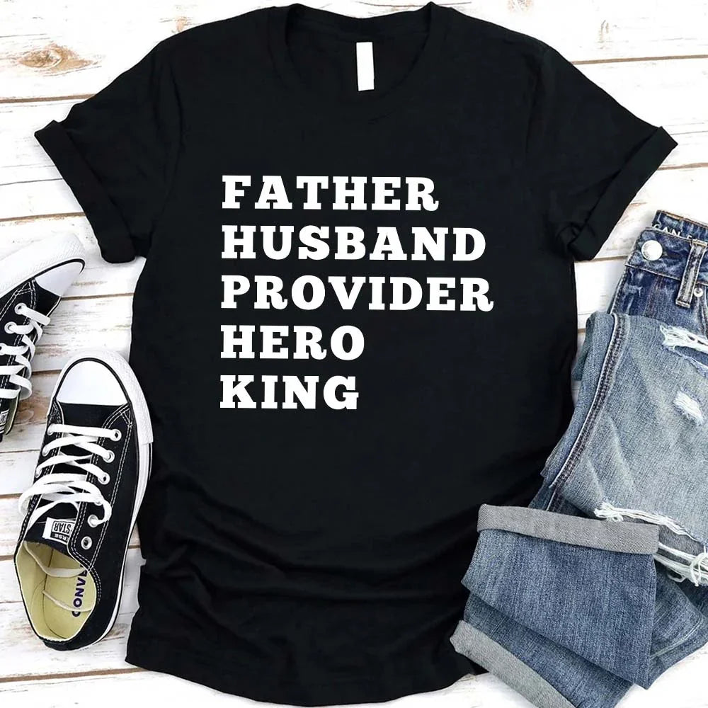 Black t-shirt with text 'Father Husband Provider Hero King' on a wooden surface with shoes and jeans.
