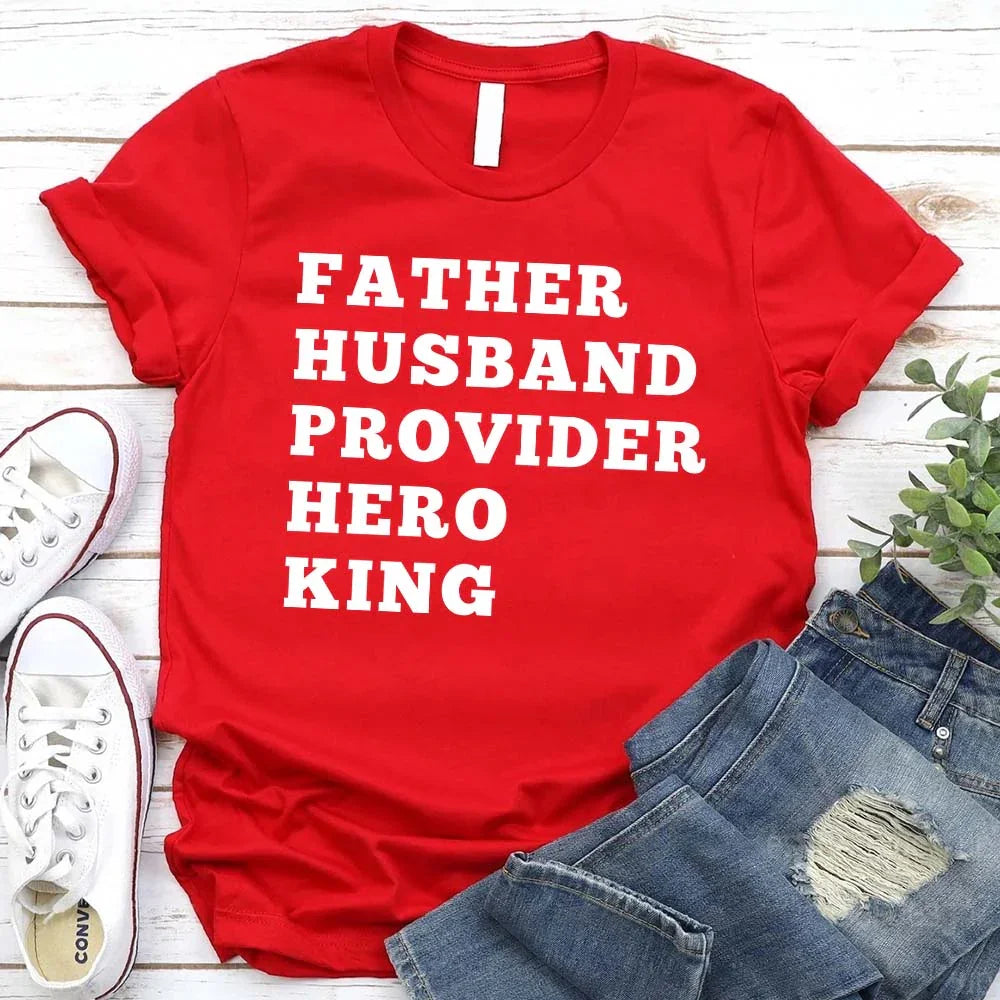 Red t-shirt with text 'FATHER HUSBAND PROVIDER HERO KING' on a wooden surface with jeans and sneakers.