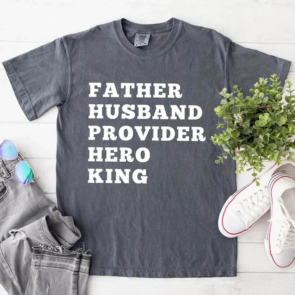 Gray t-shirt with text 'FATHER HUSBAND PROVIDER HERO KING' on a white surface with shoes and a plant.