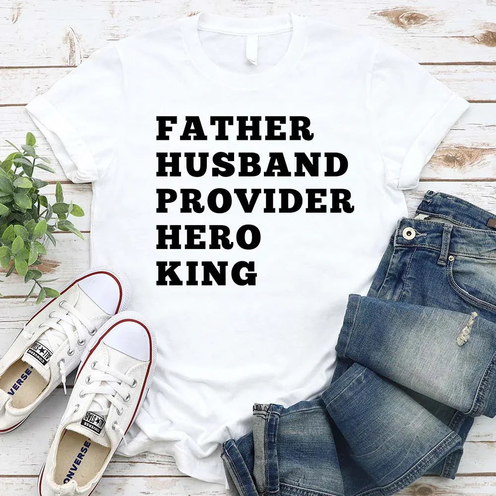 White t-shirt with text 'FATHER HUSBAND PROVIDER HERO KING' on a wooden surface with jeans and shoes.