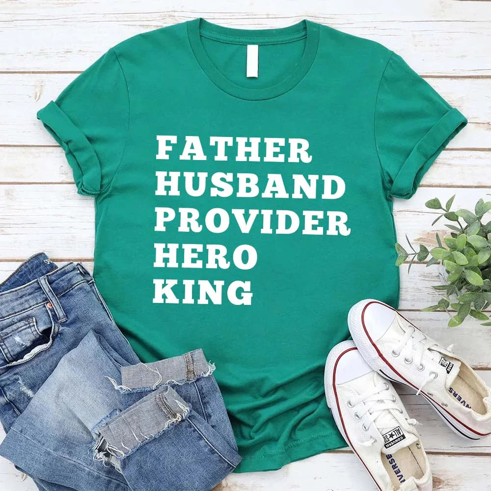 Green t-shirt with text 'FATHER HUSBAND PROVIDER HERO KING' on a wooden surface with jeans and shoes.