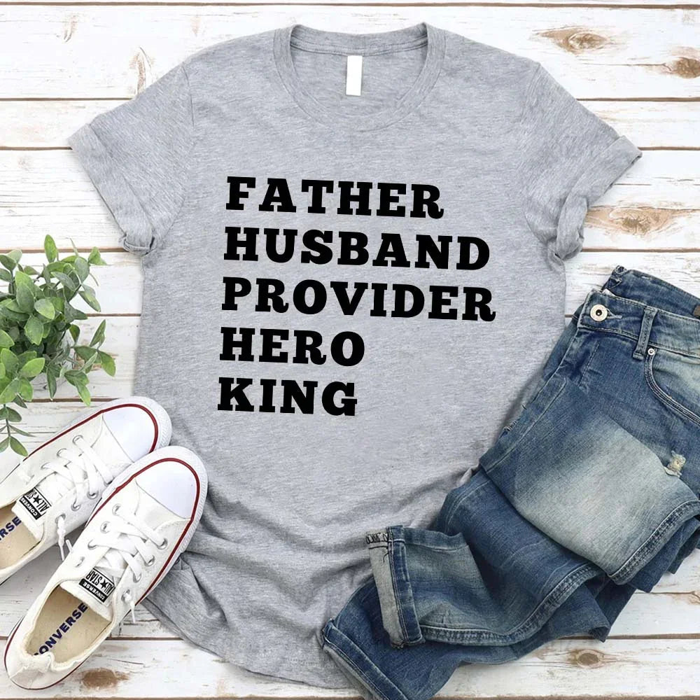 Gray t-shirt with 'FATHER HUSBAND PROVIDER HERO KING' text, paired with jeans and sneakers on a wooden surface.