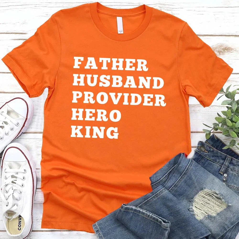 Orange t-shirt with text 'FATHER HUSBAND PROVIDER HERO KING' on a wooden surface with jeans and sneakers.